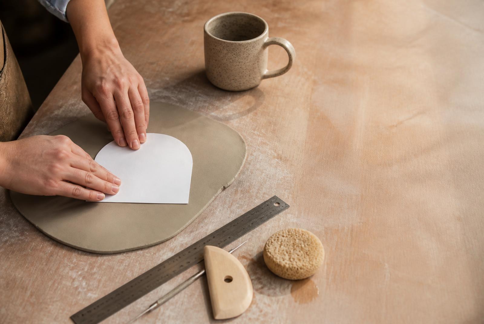 A maker transferring a paper pattern onto a rolled clay slab on a daylit pottery workbench